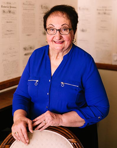 Victoria Sportelli smiles brightly at the camera, wearing a royal blue shirt and is standing behind a chair where her hands rest on its back wooden frame.